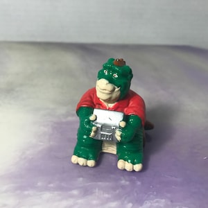 Może przedstawiać: Zielona i czerwona plastikowa figurka dinozaura trzymająca srebrny, plastikowy magnetofon. Dinozaur ma brązowy kapelusz na głowie.