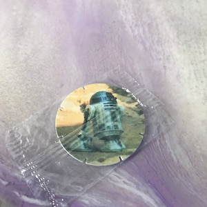 Può includere: Un gettone rotondo di plastica bianca con un'immagine di R2-D2, un robot del franchise di Star Wars, su uno sfondo desertico.