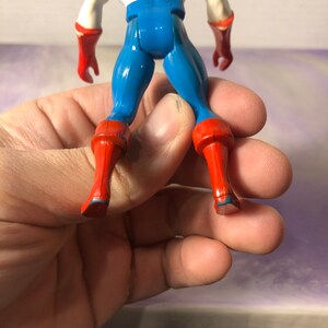 May include: A Captain America action figure, viewed from the back, showcasing a blue and white torso, blue legs, and red boots and gloves. The figure is held in a hand, with the focus on the toy's details.