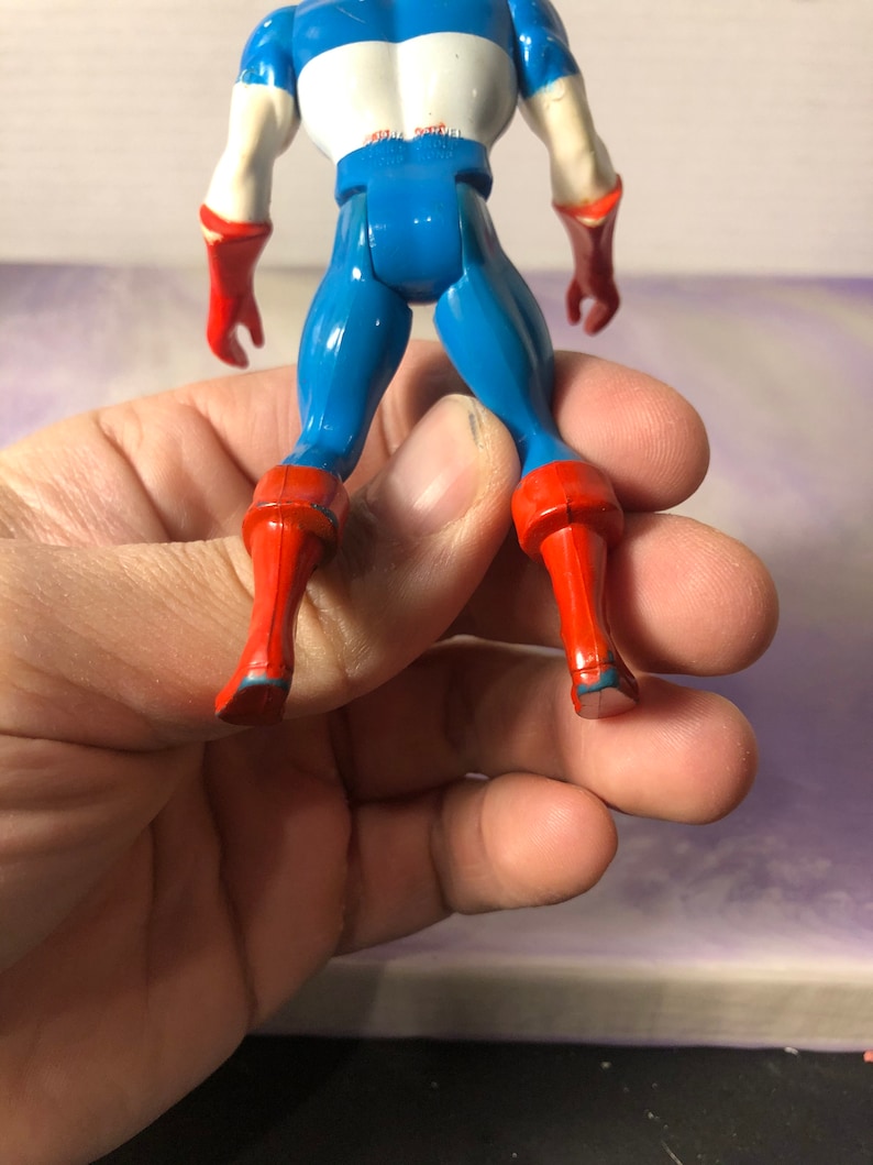 May include: A Captain America action figure, viewed from the back, showcasing a blue and white torso, blue legs, and red boots and gloves. The figure is held in a hand, with the focus on the toy's details.