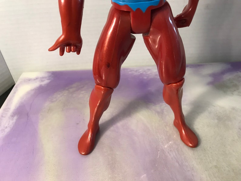 Puede incluir: Primer plano de las piernas de una figura de acci&oacute;n de pl&aacute;stico rojo. La figura tiene un torso rojo y azul y est&aacute; posada con una pierna doblada en la rodilla.