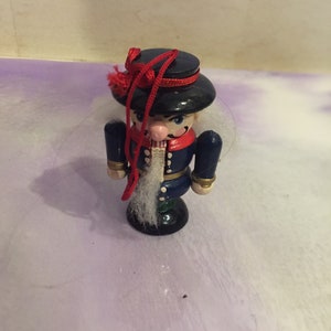 May include: A wooden nutcracker ornament with a black hat, blue uniform, and white beard. The ornament has a red ribbon tied around the hat.