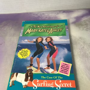 Può includere: Una copertina di un libro con due ragazze in muta da surf che cavalcano un'onda. Il titolo del libro è "The Case Of The Surfing Secret" e fa parte della serie "The New Adventures of Mary-Kate & Ashley".