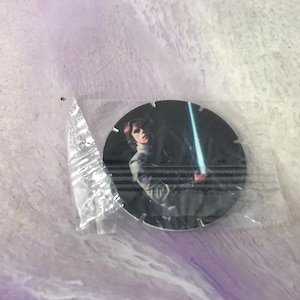 Può includere: Un oggetto da collezione rotondo, in bianco e nero, con un personaggio di Star Wars che tiene una spada laser. Il personaggio indossa una giacca scura e ha i capelli scuri.