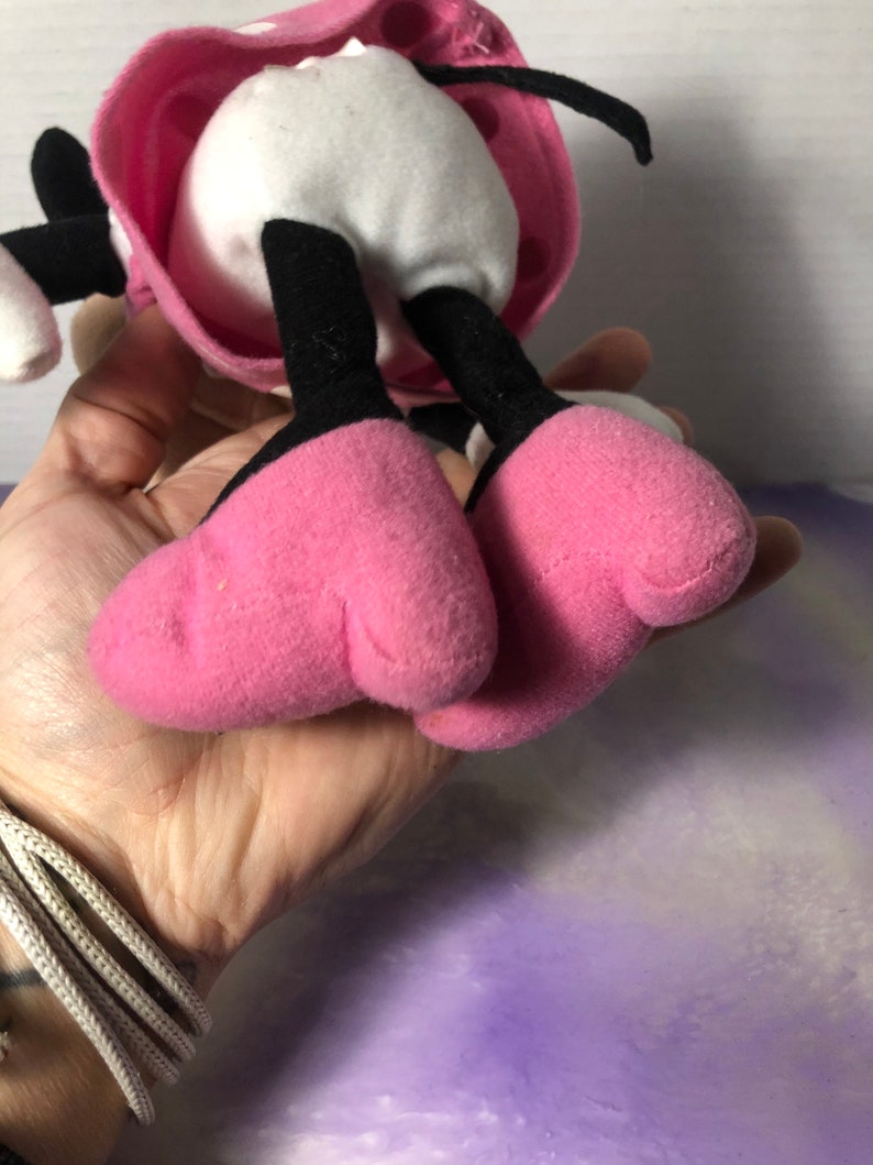 K&ouml;nnte beinhalten: Rosa und schwarzes Pl&uuml;schtier mit wei&szlig;em K&ouml;rper und schwarzen Beinen. Das Spielzeug tr&auml;gt ein rosa Kleid mit wei&szlig;en Punkten und einer schwarzen Schleife.