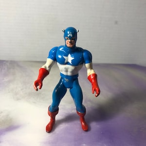 May include: A Captain America action figure in a standing pose. The figure is primarily blue, white, and red, with a white star on the chest. It has a blue helmet with the letter "A" on the forehead and red gloves and boots.