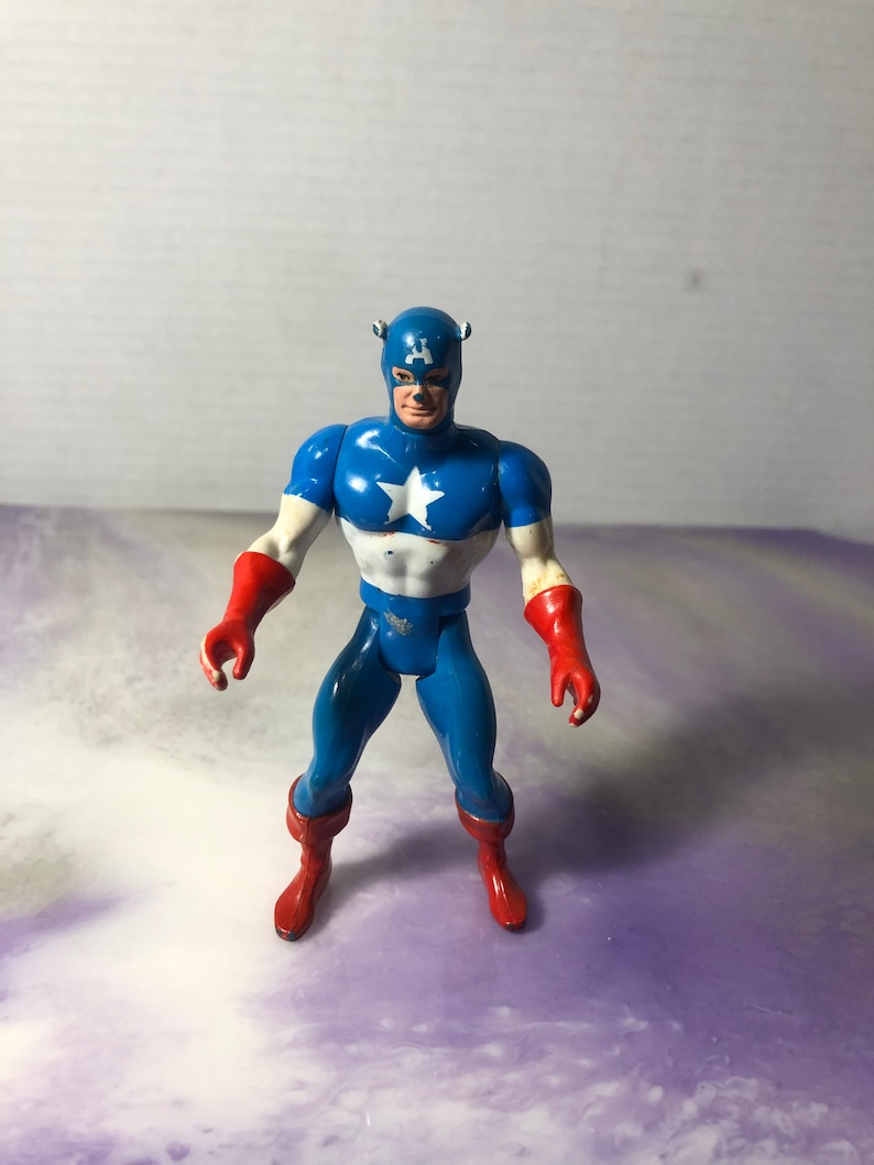 May include: A Captain America action figure in a standing pose. The figure is primarily blue, white, and red, with a white star on the chest. It has a blue helmet with the letter "A" on the forehead and red gloves and boots.