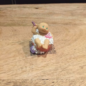 May include: A small figurine of a baby in a bathtub with a purple washcloth and a pink and white striped towel. The baby has green eyes and a pink and white striped bathing suit.