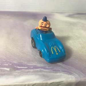 May include: A blue toy car with a yellow M logo on the side. A toy figure of a man with a burger head is driving the car.