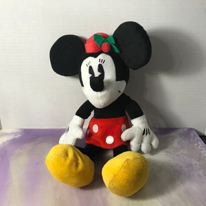May include: A plush toy of Minnie Mouse wearing a red and green holly hat. She is sitting with her legs crossed and has a red skirt with white polka dots.