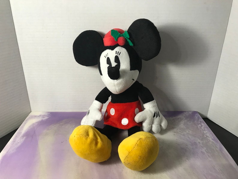 May include: A plush toy of Minnie Mouse wearing a red and green holly hat. She is sitting with her legs crossed and has a red skirt with white polka dots.