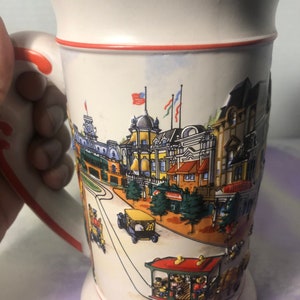 K&ouml;nnte beinhalten: Ein wei&szlig;er Keramik-Bierkrug mit rotem Rand und Henkel. Der Krug zeigt eine farbenfrohe Illustration der Main Street, U.S.A. in Walt Disney World. Die Illustration umfasst Geb&auml;ude, B&auml;ume und eine Stra&szlig;enbahn mit Disney-Charakteren.