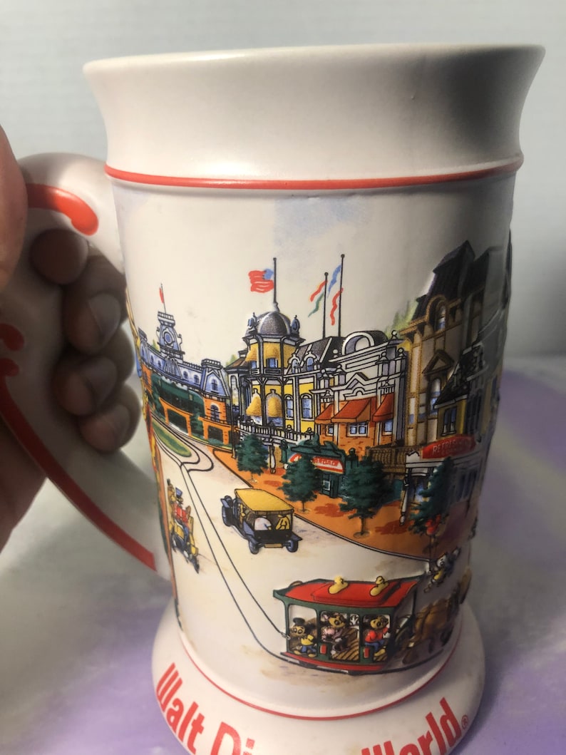 K&ouml;nnte beinhalten: Ein wei&szlig;er Keramik-Bierkrug mit rotem Rand und Henkel. Der Krug zeigt eine farbenfrohe Illustration der Main Street, U.S.A. in Walt Disney World. Die Illustration umfasst Geb&auml;ude, B&auml;ume und eine Stra&szlig;enbahn mit Disney-Charakteren.