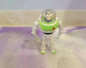 May include: A white and green plastic action figure of Buzz Lightyear from the Toy Story franchise. The figure has a clear plastic helmet with a painted face and a green and white spacesuit.