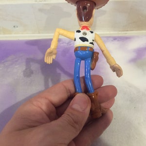 May include: A plastic toy figure of Woody from the Toy Story franchise. Woody is wearing his signature brown cowboy hat, blue jeans, and a brown vest with a white and black cow pattern. He is holding a brown holster with a gun on his right hip.