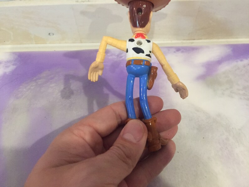 May include: A plastic toy figure of Woody from the Toy Story franchise. Woody is wearing his signature brown cowboy hat, blue jeans, and a brown vest with a white and black cow pattern. He is holding a brown holster with a gun on his right hip.