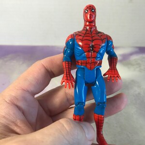 May include: A vintage action figure of Spider-Man, wearing a red and blue suit with a spider logo on the chest. The figure has articulated arms and legs, and is posed in a standing position.