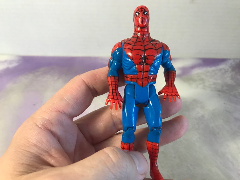 May include: A vintage action figure of Spider-Man, wearing a red and blue suit with a spider logo on the chest. The figure has articulated arms and legs, and is posed in a standing position.