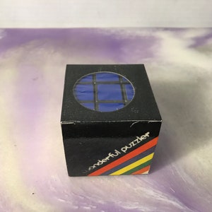 May include: A black box with a clear window showing a blue Rubik's Cube. The box has the text "wonderful puzzler" printed on the front.