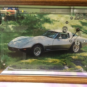 May include: A silver Corvette sports car is featured on a clock face with black hands and white numbers. The car is on a green grassy background.
