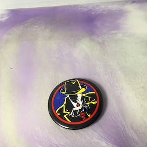 Może przedstawiać: Okrągła, czarna przypinka z ilustracją w stylu pop-art przedstawiającą mężczyznę w kapeluszu i garniturze, spoglądającego na zegarek. Projekt zawiera żółte, czerwone, niebieskie i białe akcenty.