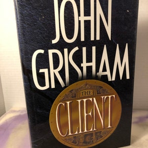 May include: A hardcover book with a black cover and gold lettering. The title of the book is "The Client" by John Grisham.