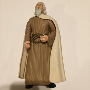 May include: A white-haired action figure wearing a brown robe and a white cloak. The figure is holding a brown beaded necklace in its right hand.