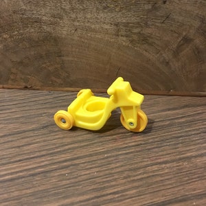 May include: A yellow plastic toy tricycle with orange wheels. The tricycle is a vintage toy.
