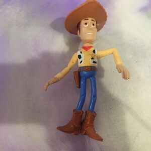 May include: A plastic toy figure of Woody from the Toy Story franchise. Woody is wearing his signature brown cowboy hat, yellow shirt with a red bandana, blue jeans, and brown boots. He has a friendly expression on his face.