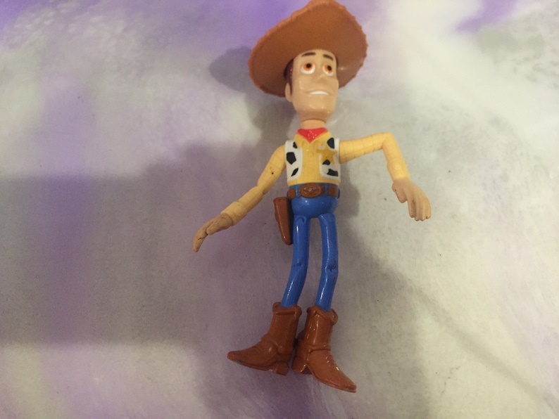 May include: A plastic toy figure of Woody from the Toy Story franchise. Woody is wearing his signature brown cowboy hat, yellow shirt with a red bandana, blue jeans, and brown boots. He has a friendly expression on his face.