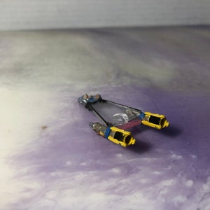 以下が含まれることがあります： 2 つのエンジンが付いた、小さく青い黄色の宇宙船のおもちゃ。宇宙船は、紫と白の背景の上にあります。
