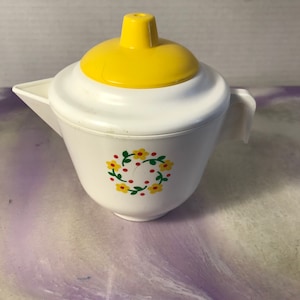 Op de afbeelding: Witte plastic theepot met een geel deksel en een bloemenmotief. De theepot heeft een handvat en een tuit.