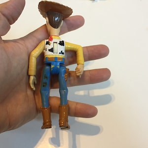 Op de afbeelding: Een plastic speelgoedfiguur van Woody uit Toy Story. Hij draagt een bruine cowboyhoed, een geel shirt met een rood vest, een blauwe spijkerbroek en bruine laarzen. Hij heeft een trekkoord op zijn rug.