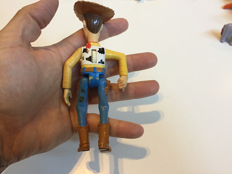 Op de afbeelding: Een plastic speelgoedfiguur van Woody uit Toy Story. Hij draagt een bruine cowboyhoed, een geel shirt met een rood vest, een blauwe spijkerbroek en bruine laarzen. Hij heeft een trekkoord op zijn rug.