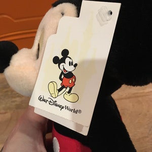 Może przedstawiać: Pluszowa zabawka Mickey Mouse w kolorze czarnym i czerwonym z białą metką z napisem "Walt Disney World&reg;" i rysunkową postacią Myszki Miki.