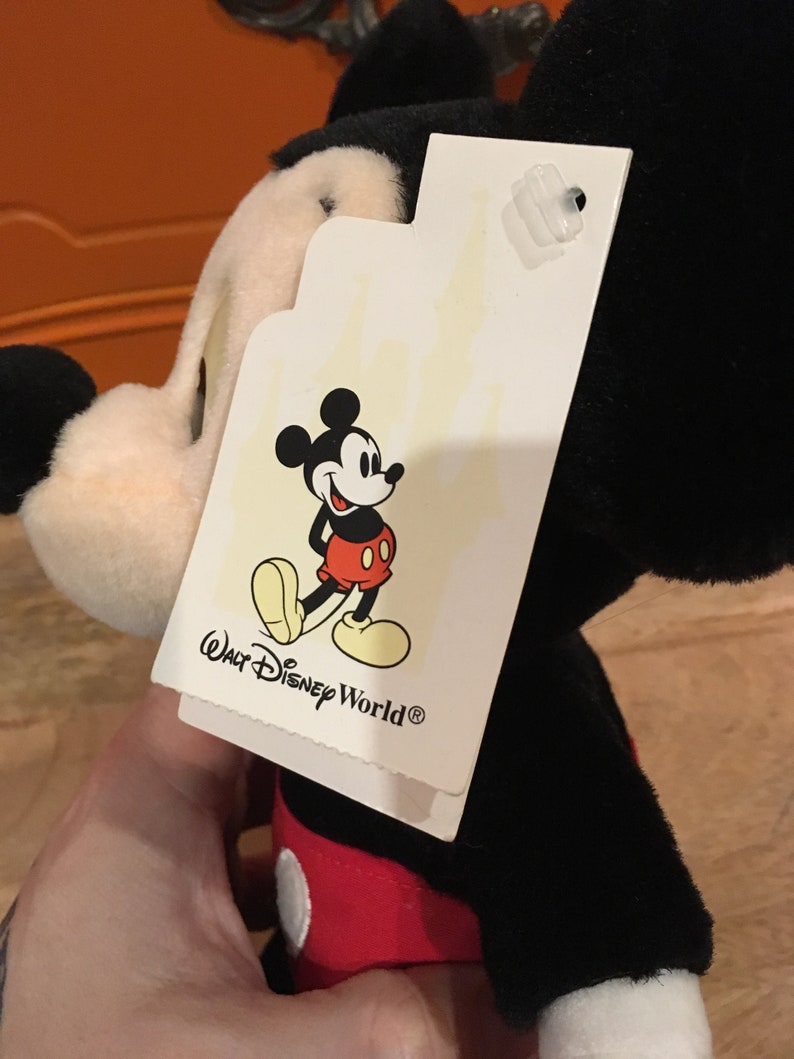 Może przedstawiać: Pluszowa zabawka Mickey Mouse w kolorze czarnym i czerwonym z białą metką z napisem "Walt Disney World&reg;" i rysunkową postacią Myszki Miki.