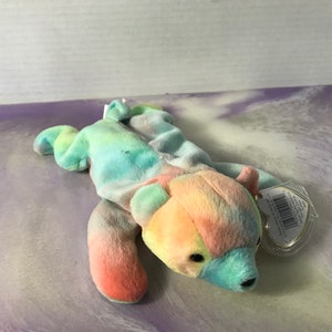 May include: A tie-dye plush bear with a rainbow colour pattern. The bear is lying on its side with its head turned to the right. The bear has a small tag attached to its ear.