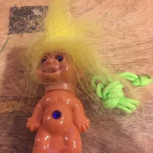 May include: A small plastic troll doll with yellow hair and a blue gem on its belly. The troll is wearing a green string necklace.