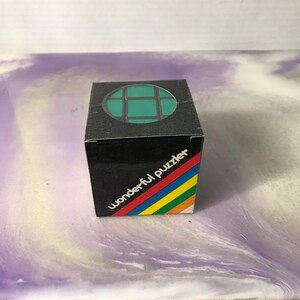 Może przedstawiać: Czarne pudełko z zielono-czarną układanką w środku. Na pudełku widnieje napis "wonderful puzzler".