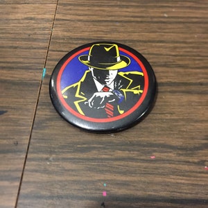 Peut inclure: Un bouton noir et jaune avec une image de dessin animé d'un homme portant un fedora et un costume. L'homme tient une arme à feu et regarde sur le côté. L'arrière-plan est bleu et rouge.