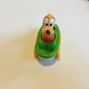 May include: A green toy boat with yellow wheels and a brown and orange cartoon dog figure in the driver's seat.