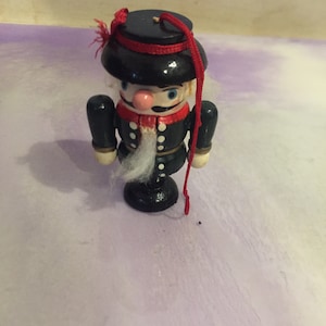 May include: A wooden nutcracker ornament with a black hat, red trim, and a white beard. The ornament is painted with blue and white details and has a red string attached.
