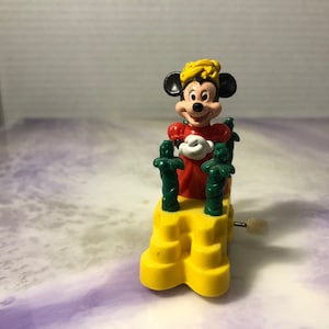 May include: A yellow wind-up toy featuring Minnie Mouse in a red dress and a yellow crown. She is surrounded by green palm trees and is standing on a yellow base.