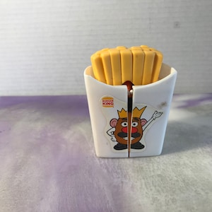 Pode incluir: Um recipiente de plástico branco com um logotipo do Burger King e um personagem do Mr. Potato Head usando uma coroa. O recipiente está cheio de batatas fritas de plástico amarelas.