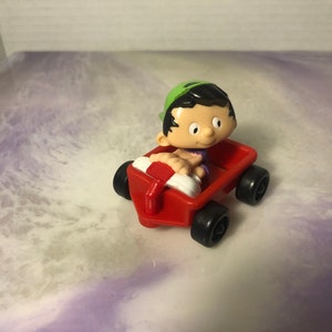 May include: A red toy wagon with a small cartoon boy figure in a green hat and purple shirt. The boy is holding a white life preserver and is sitting in the wagon.