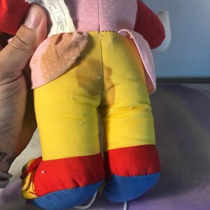 Op de afbeelding: Een pluche speelgoed met een roze, rode en gele outfit. Het speelgoed heeft een wit touwtje aan zijn voeten. Het speelgoed is gemaakt van zacht materiaal en heeft een versleten uiterlijk.