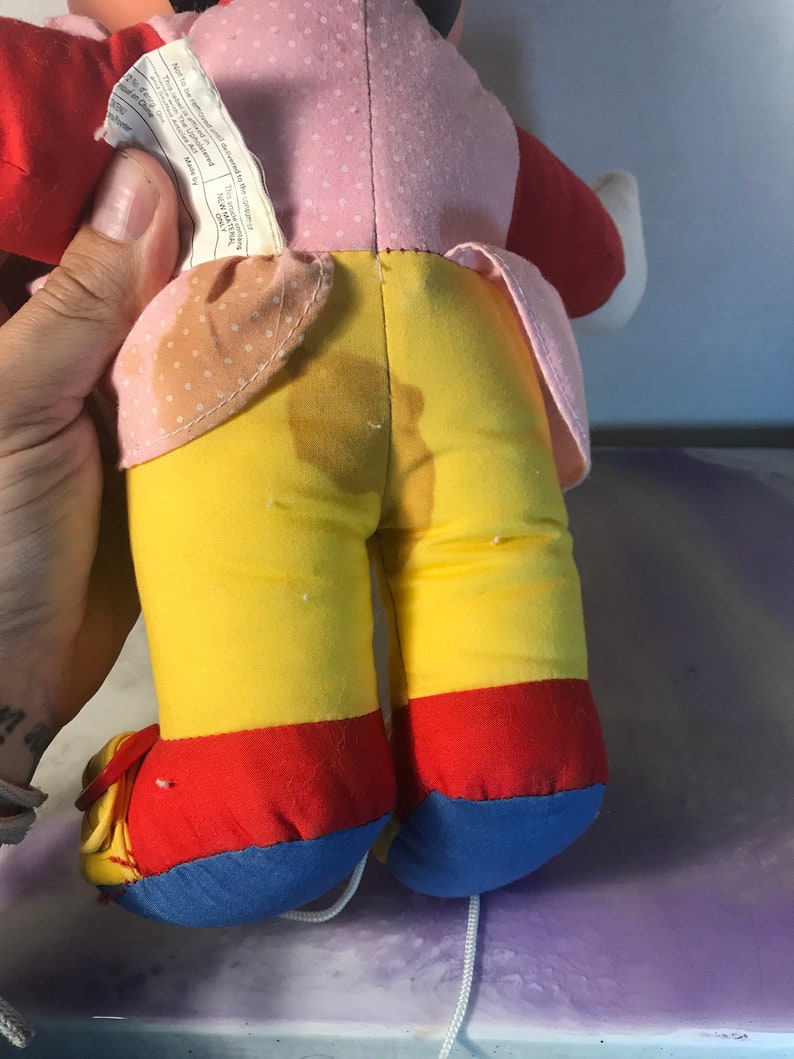 Op de afbeelding: Een pluche speelgoed met een roze, rode en gele outfit. Het speelgoed heeft een wit touwtje aan zijn voeten. Het speelgoed is gemaakt van zacht materiaal en heeft een versleten uiterlijk.