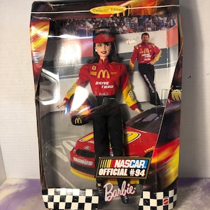 May include: A Barbie doll dressed in a red and yellow racing suit with the McDonald's logo. The doll is holding a McDonald's hat and is standing in front of a red race car with the number 94 on it. The box says "NASCAR Official #94 Barbie".