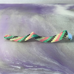 May include: A close-up of a multi-colored woven bracelet with pink, green, black, and white stripes. The bracelet is made of small beads and is lying on a purple and white surface.