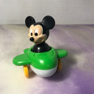 May include: A green and white toy aeroplane with Mickey Mouse in the cockpit. The aeroplane has yellow wheels and Mickey Mouse has black ears and a black nose.